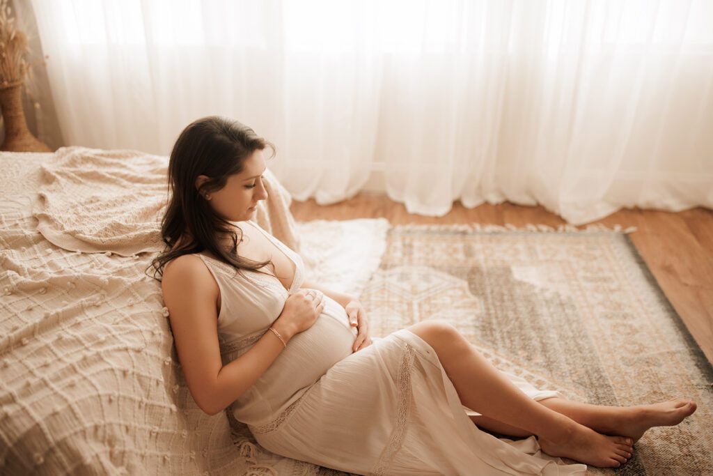 what to wear for your maternity photo session - pregnant mama in cream lace dress leaning against a bed