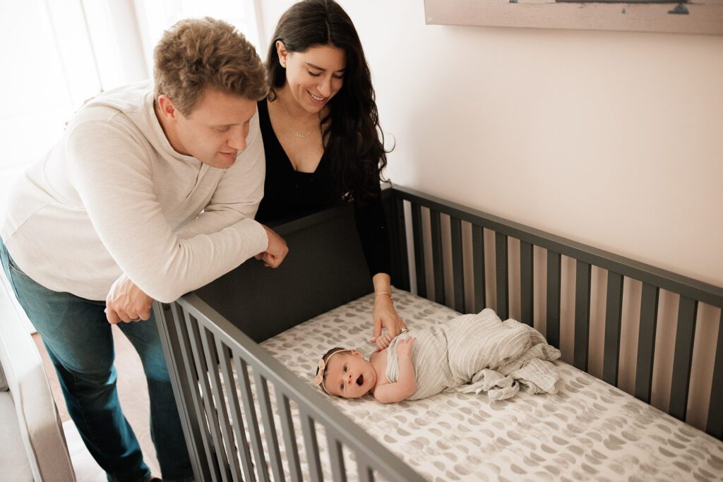 Which type of newborn photo session is best - parents looking at baby girl laying in her crib in nursery in home lifestyle