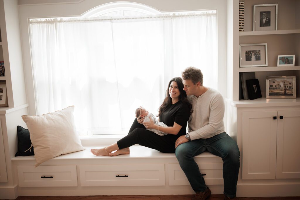 Which type of newborn photo session is best - parents sitting on window bench with baby girl in home lifestyle photos