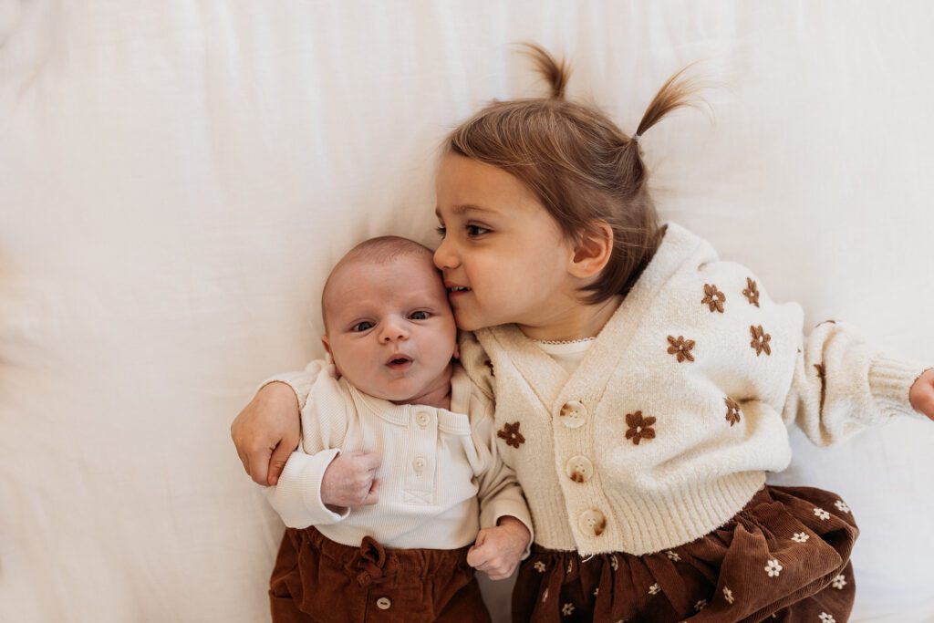 How old should my baby be for newborn photos? - big sister toddler laying on bed with flower sweater cream and brown and little baby newborn brother kissing head lifestyle family session in home