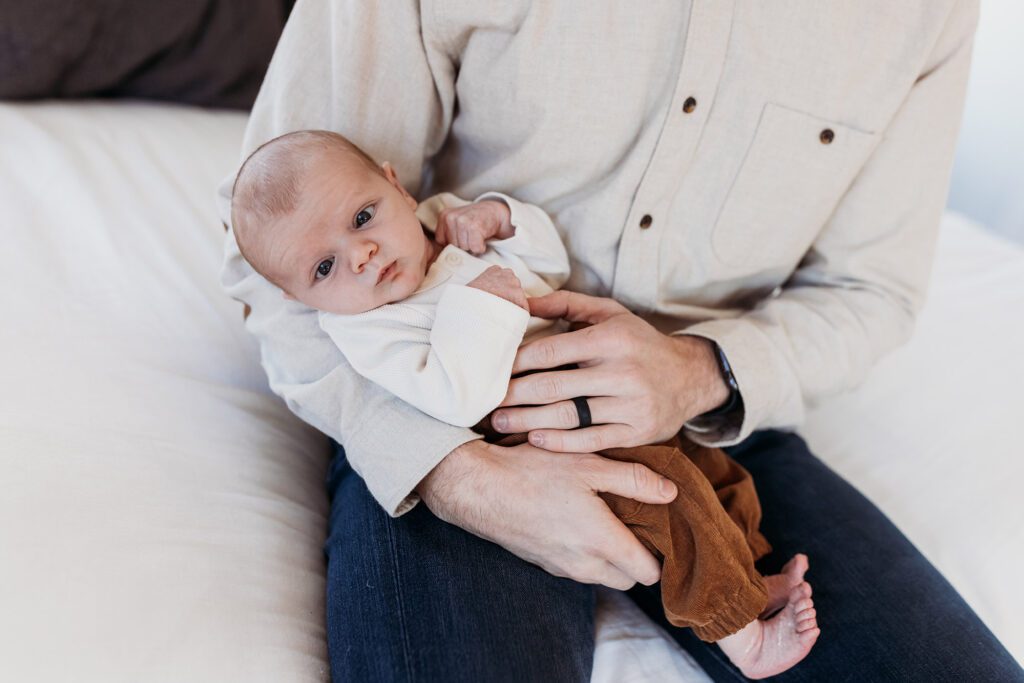 How old should my baby be for newborn photos? - dad in cream button down and jeans sitting on a bed with baby in his arms in brown pants and white top
