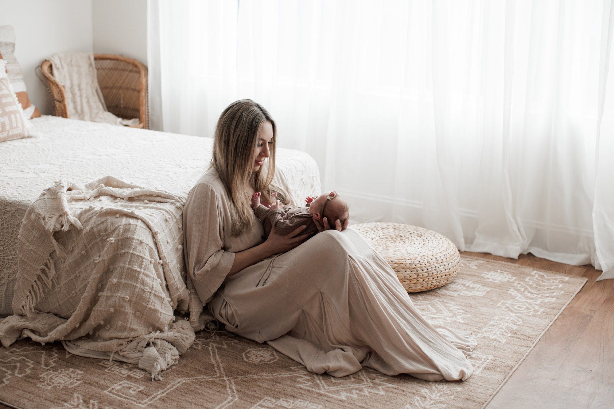 How to choose the right newborn photographer for your family - mom sitting on the floor with a baby in her lap in front of a bed with a white window
