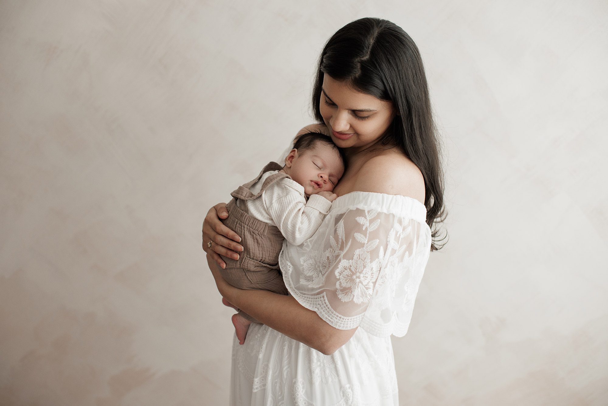 When should I book my newborn photos? - mom holding baby on her chest with plain backdrop ad white dress